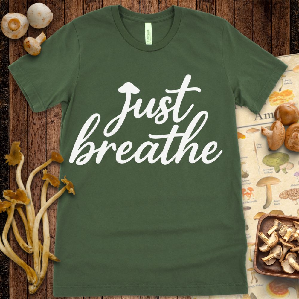 Black T-shirt design with cursive white text that says "Just breathe" with a small mushroom cap as the dot on the "J"