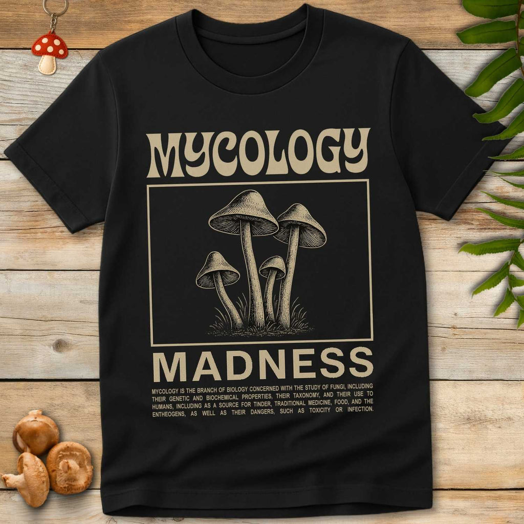 Black t-shirt with 'Mycology Madness' graphic on a wooden surface with mushrooms and leaves.