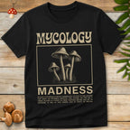 Black t-shirt with 'Mycology Madness' graphic on a wooden surface with mushrooms and leaves.