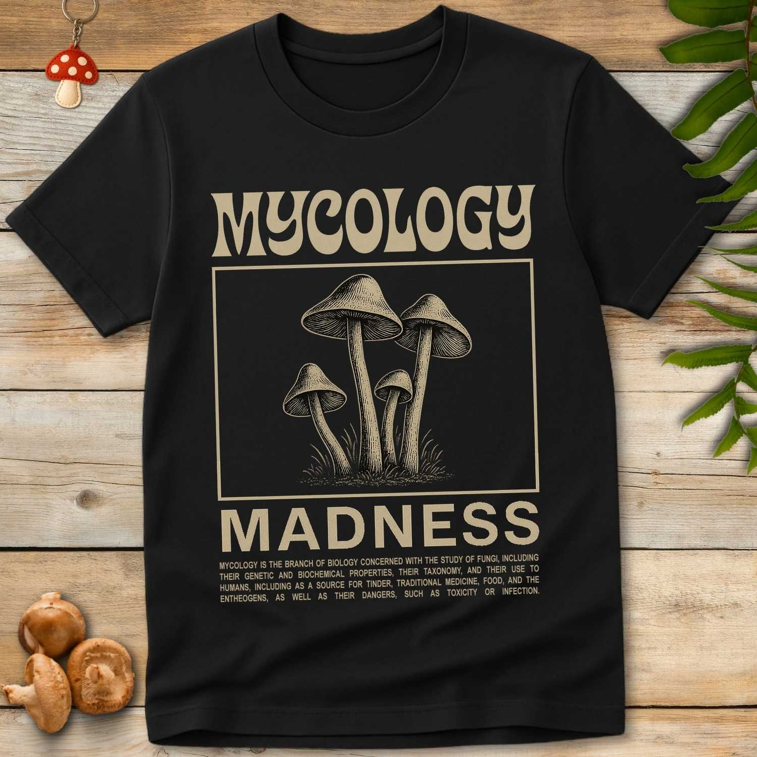 Black t-shirt with 'Mycology Madness' graphic on a wooden surface with mushrooms and leaves.