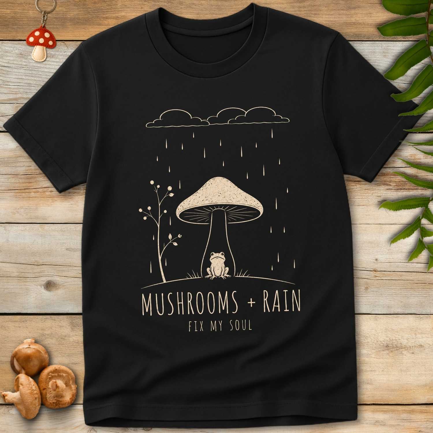 Mushroom Rain Tee featuring a frog sitting under a giant mushroom during rainfall with cozy woodland cottagecore artwork.