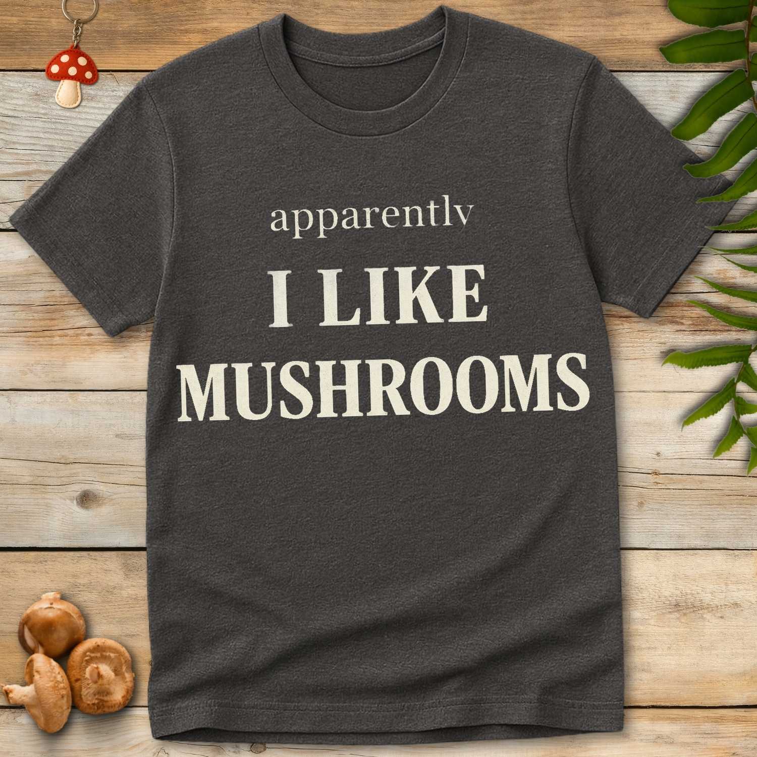 Minimalist typographic t-shirt design reading “apparently, I like mushrooms” in bold vintage-style white lettering on a grey background.