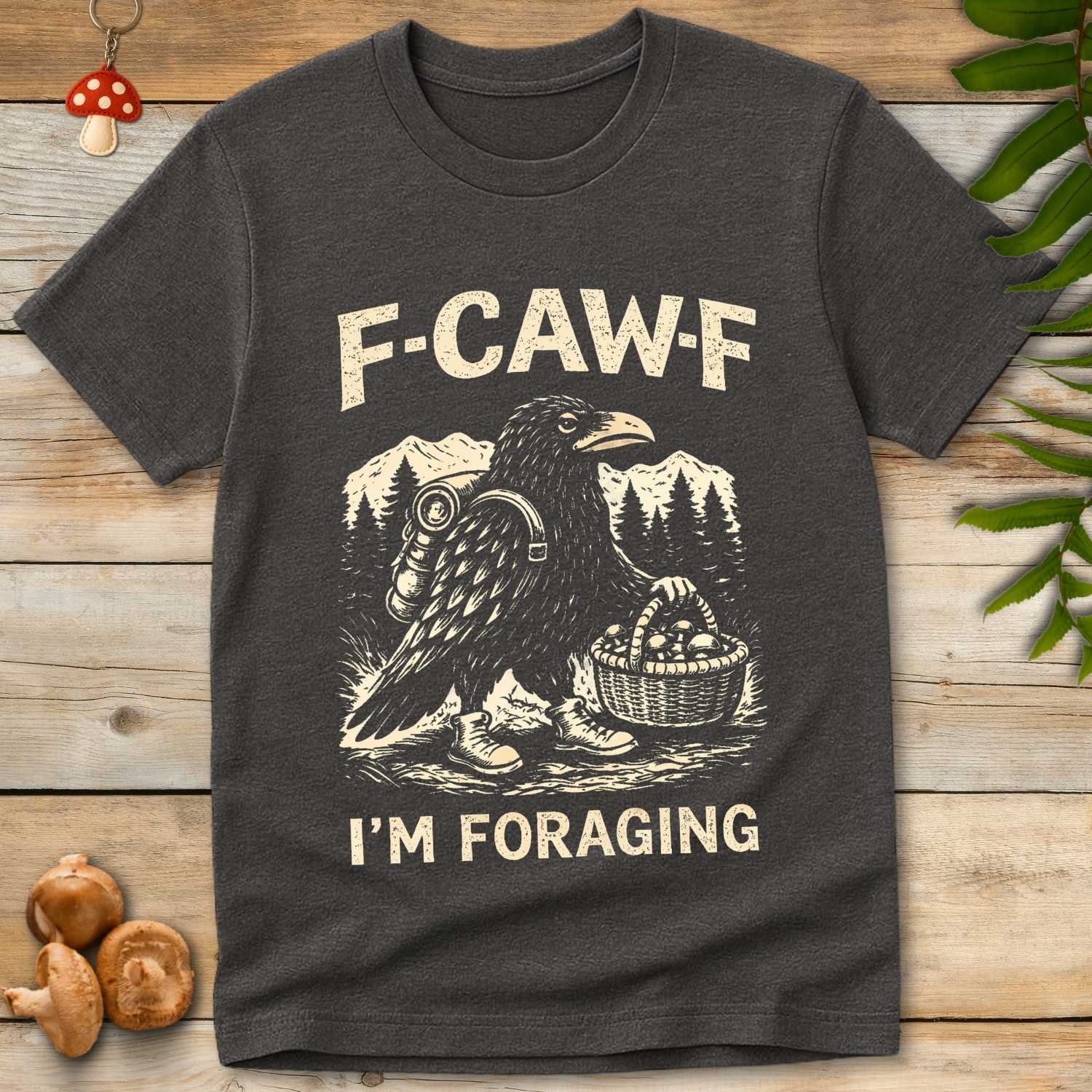 T-shirt graphic of a crow wearing boots and carrying a mushroom basket with the phrase “F-CAW-F I’m Foraging” over a forest and mountain backdrop