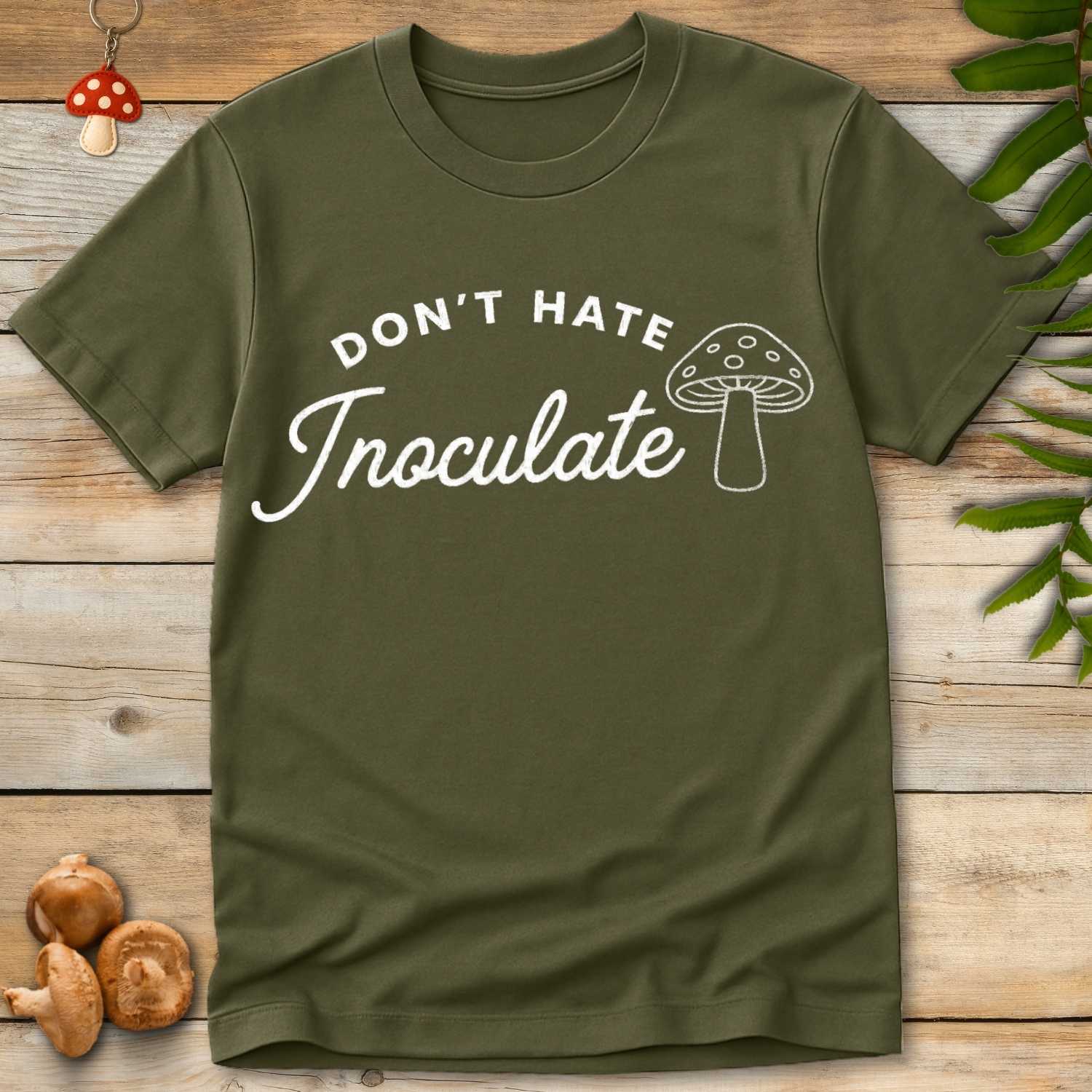 Green T-shirt with white text “Don’t Hate Inoculate” in curved bold and script fonts, with line-art mushroom illustration