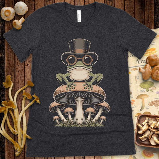 Green frog in top hat and goggles sitting on a cluster of mushrooms on a graphic tee