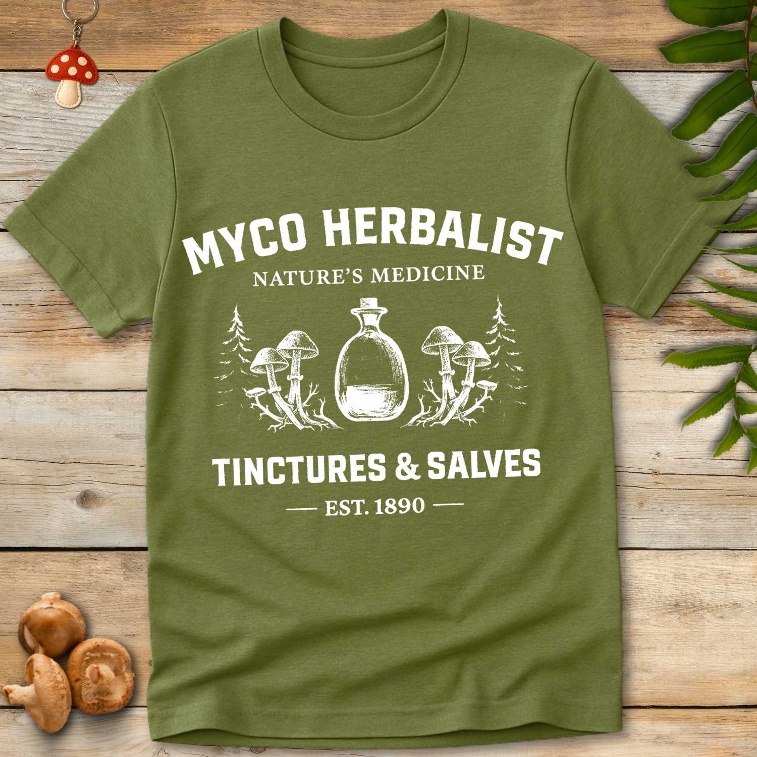 Green T-shirt with vintage white graphic reading “Myco Herbalist – Nature’s Medicine – Tinctures & Salves – Est. 1890” with mushroom and bottle illustrations