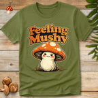 Cute smiling cartoon mushroom with orange spotted cap and vintage “Feeling Mushy” lettering on a green background, whimsical fungi themed t-shirt design.