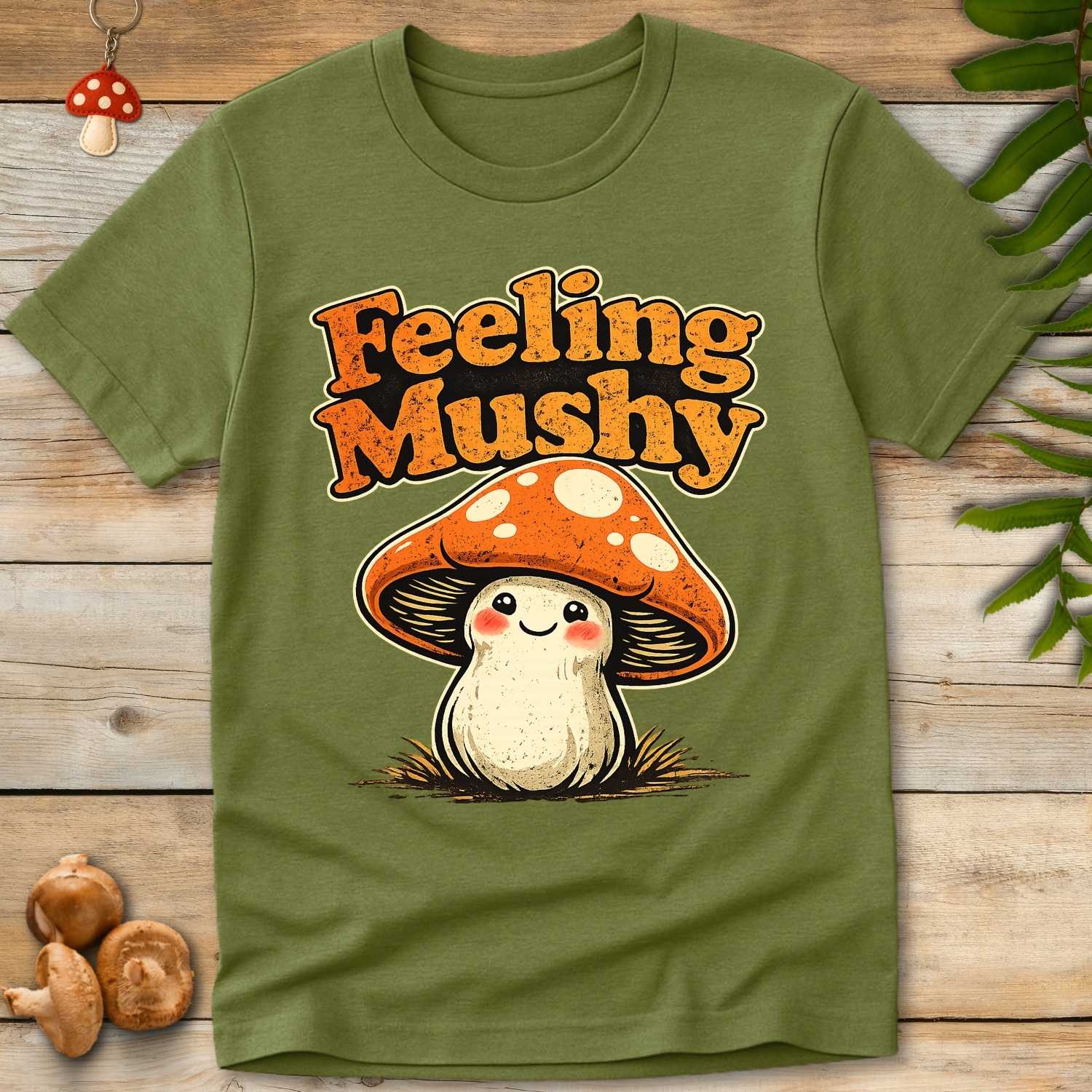 Cute smiling cartoon mushroom with orange spotted cap and vintage “Feeling Mushy” lettering on a green background, whimsical fungi themed t-shirt design.