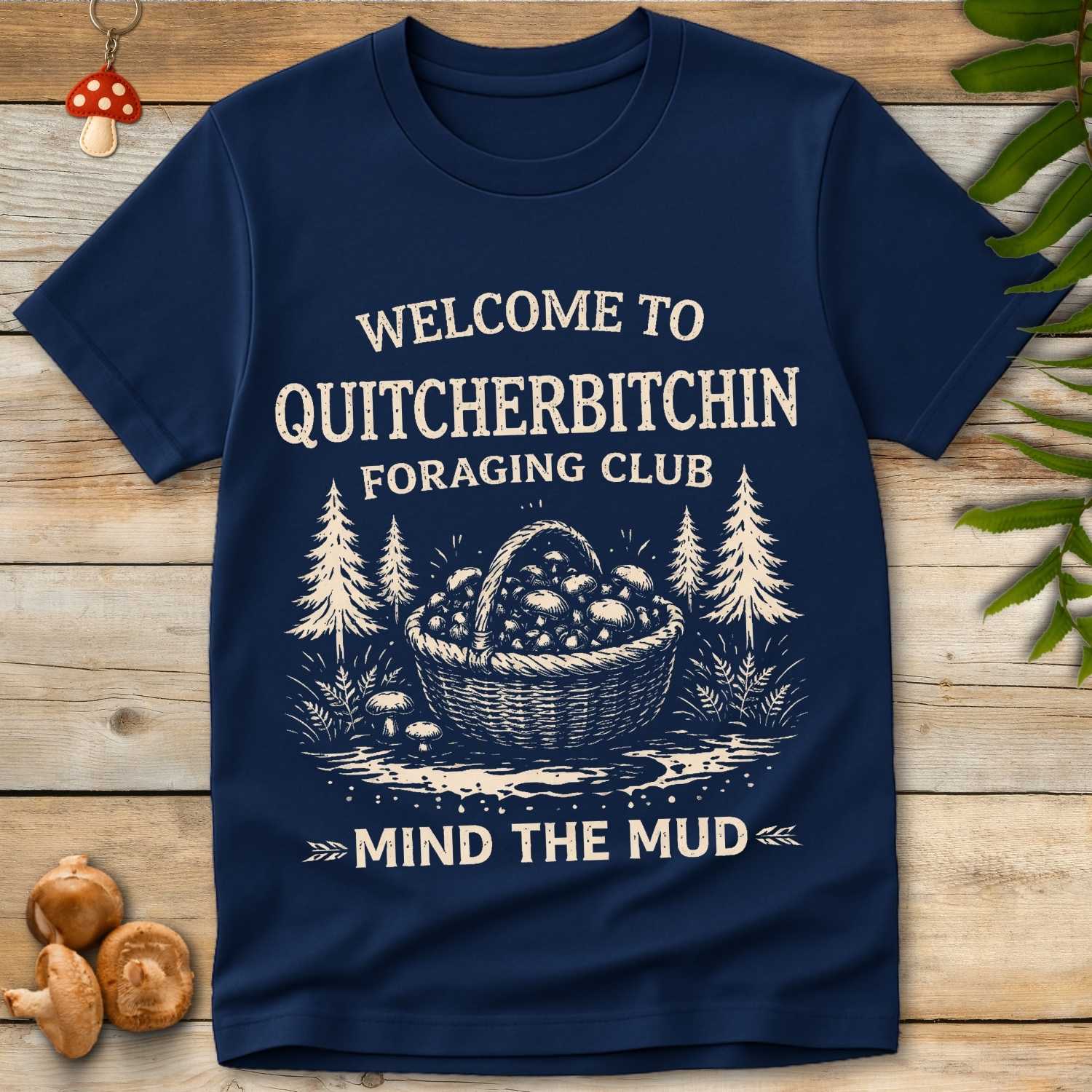 Vintage graphic t-shirt design with a basket full of mushrooms, pine trees, and bold text saying “Quitcherbitchin Foraging Club – Mind the Mud”