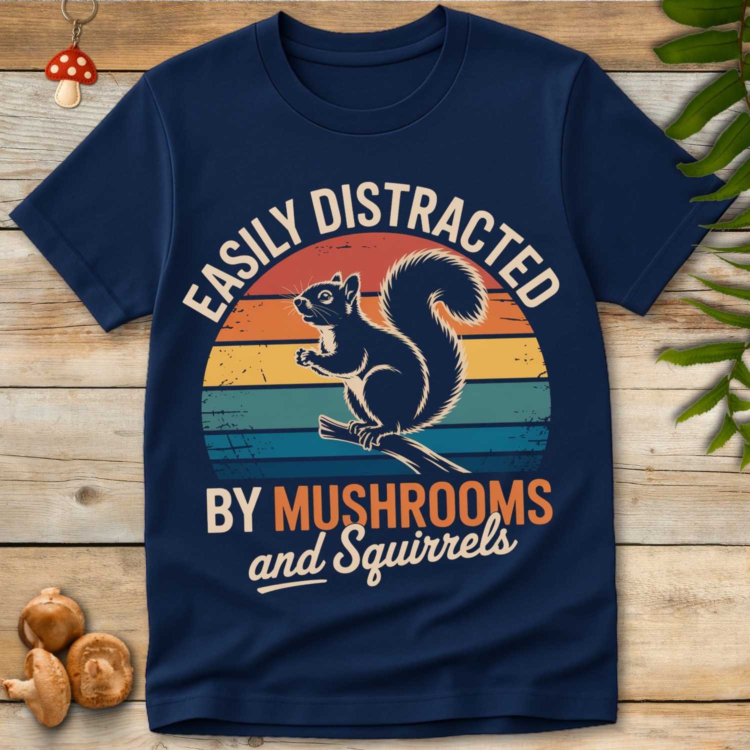 Navy blue t-shirt with a graphic of a squirrel and text 'Easily Distracted by Mushrooms and Squirrels' on a wooden surface.